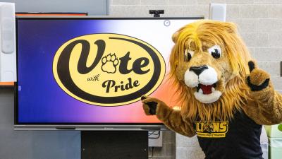 Roary posing in front of a TV that says "Vote with Pride"