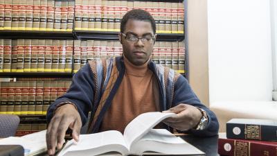 A student reads a legal text book with bookshelves filled with legal books behind him.