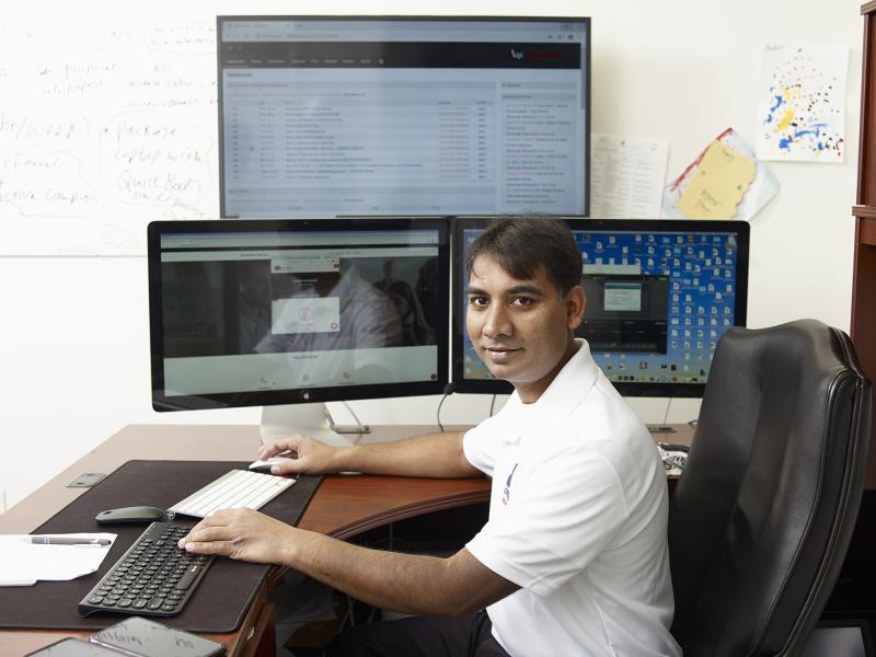 Man looks at the camera, seated at a desk with three computer screens and two keyboards.